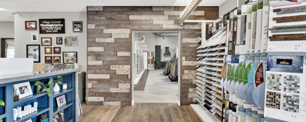 Interior of the Village Flooring & Renovation showroom hallway with wood-look flooring, stone-texture wall panels, framed photos on the left, and shelves of brochures and tile samples on the right leading to a corridor.